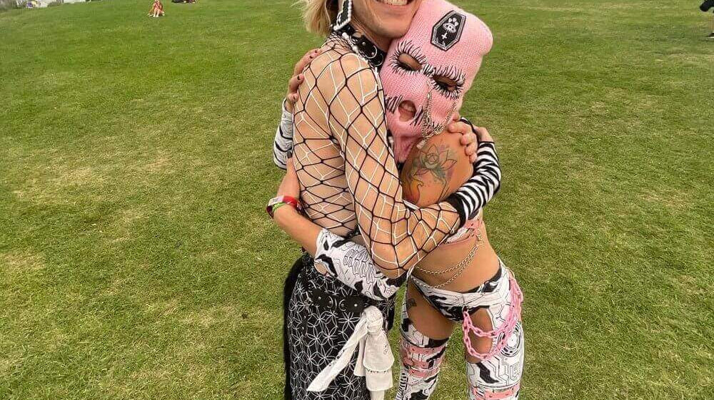 Two festival-goers embracing, wearing colorful, bold outfits, standing on a grassy field with trees in the background.