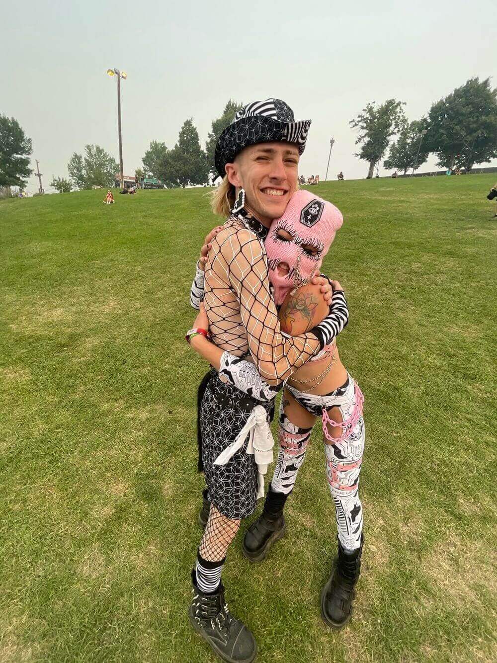 Two festival-goers embracing, wearing colorful, bold outfits, standing on a grassy field with trees in the background.