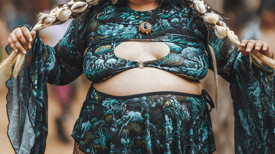A smiling woman in the forest wears plus-size rave outfits, featuring a vibrant two-piece set with green and black patterns, accessorized with fishnet stockings and braided hair, embracing the festival vibe