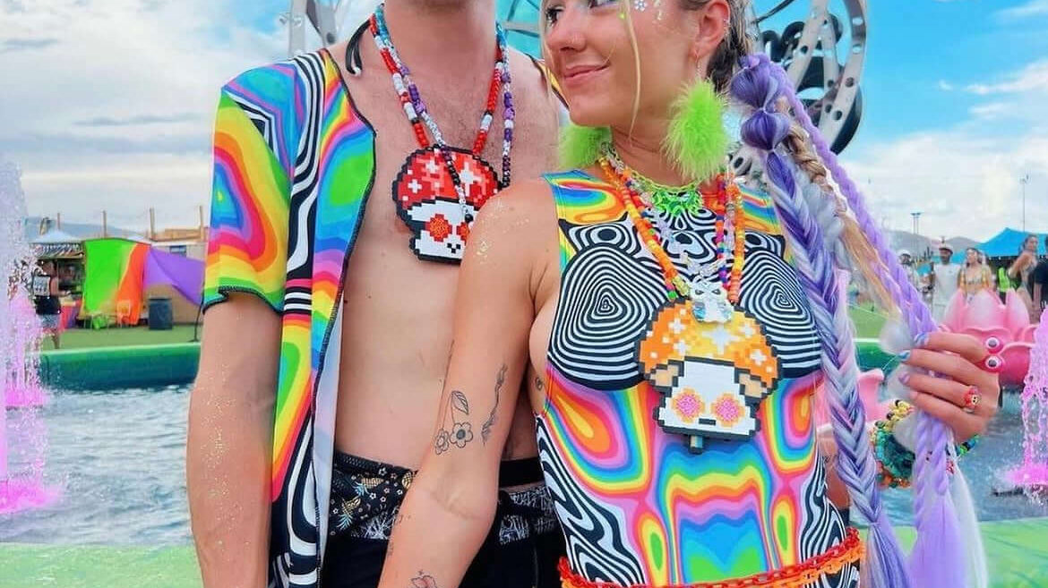A couple poses at a festival, both wearing colorful psychedelic-patterned rave outfits. She wears a fishnet bodysuit with accessories, and he wears a matching shirt and hat