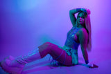 Model in vibrant rave outfit with glossy knee-high boots, posing against a colorful illuminated background.