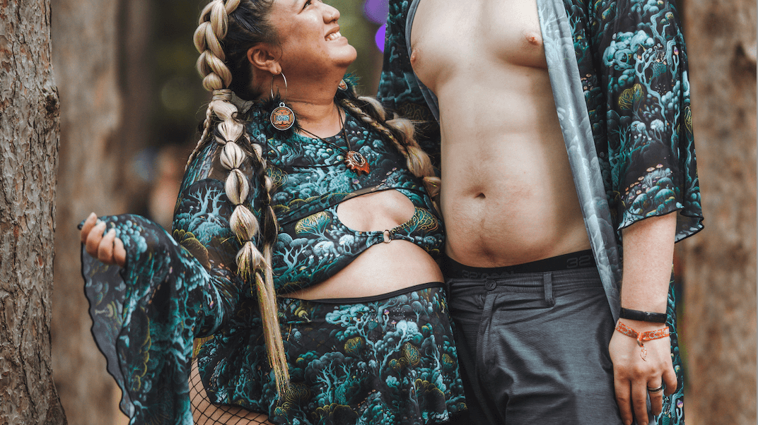 A joyful couple dressed in matching festival outfits, the woman in a plus-size green and black patterned two-piece set with fishnet stockings, and the man in matching shorts and an open robe, sharing a happy moment in the forest.