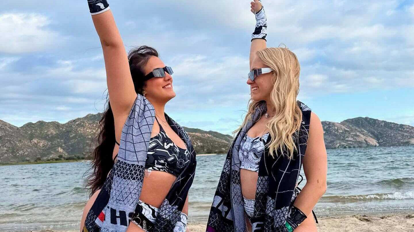 Two women in rave outfits posing on the beach, celebrating friendship with a scenic backdrop.