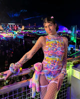 raver in a bright rainbow outfit with matching fluffy rainbow bear stands at the top of the bleachers at edc in front of the stages