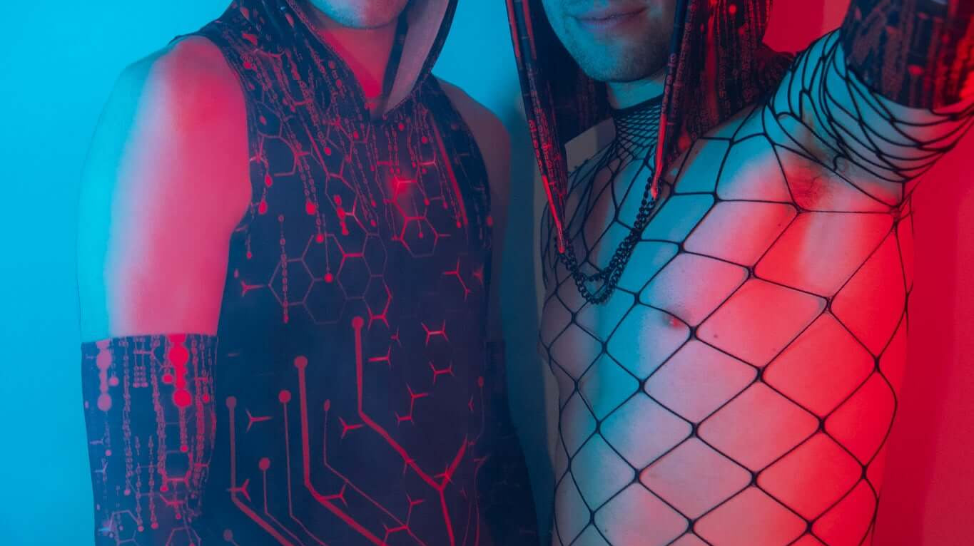 Two men wearing stylish rave gear, including a hooded sleeveless top with a futuristic red and black circuit design and a fishnet top, standing in dramatic blue and red lighting.
