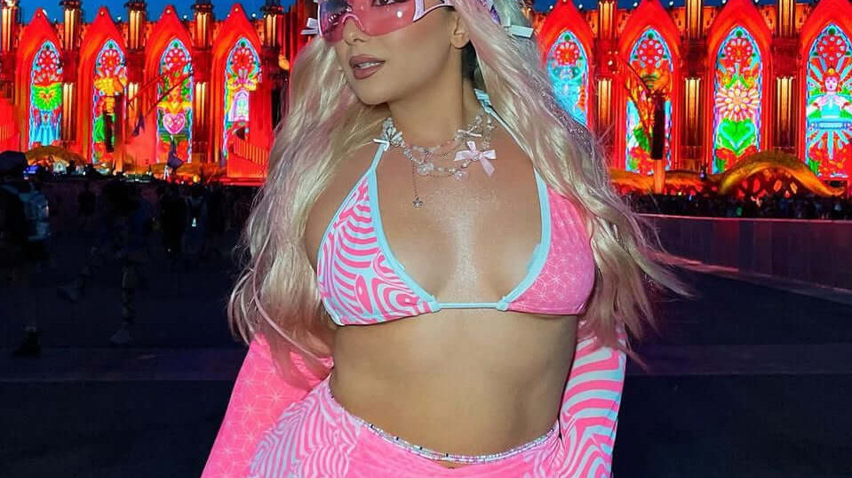 A festival-goer in a vibrant women's rave outfit poses confidently. She wears a pink patterned bikini set with matching accessories, thigh-high socks, and sunglasses, set against a colorful festival backdrop at night