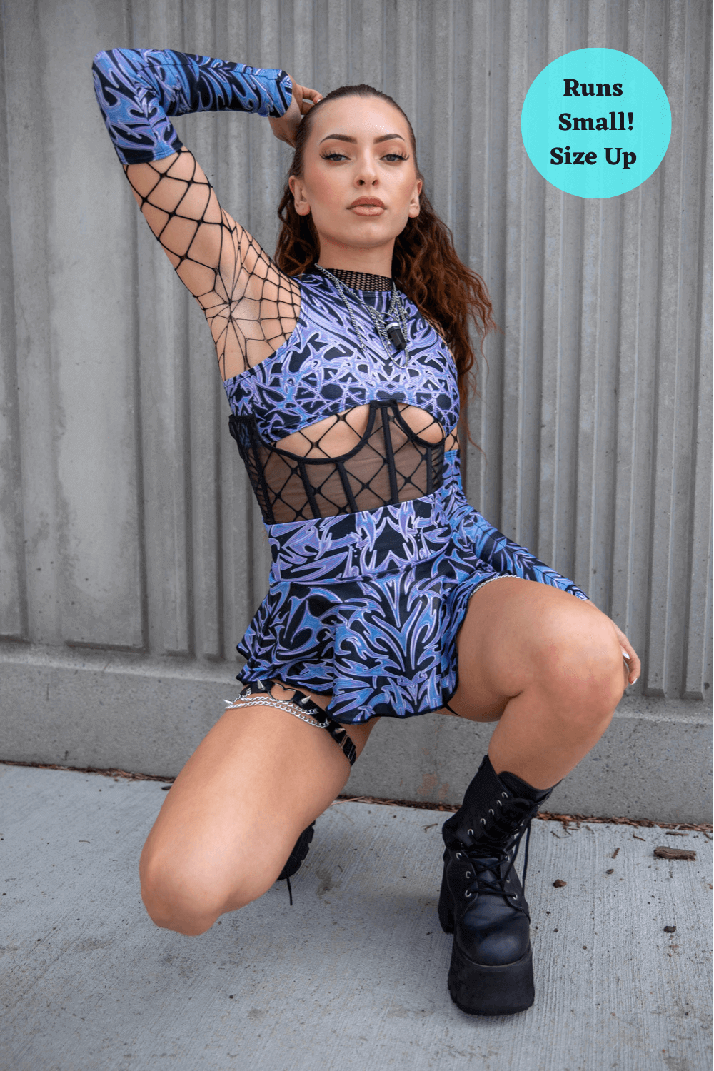 Model in edgy rave outfit featuring blue floral patterns and fishnet accents, posing against a textured wall.