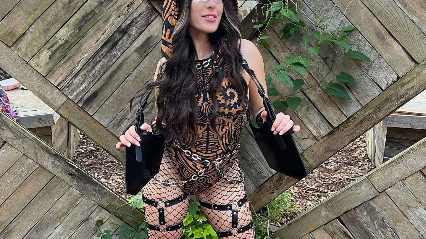 A woman wearing a hooded bodysuit, fishnet stockings, and boots poses in front of a wooden structure with greenery, showcasing stylish rave accessories.
