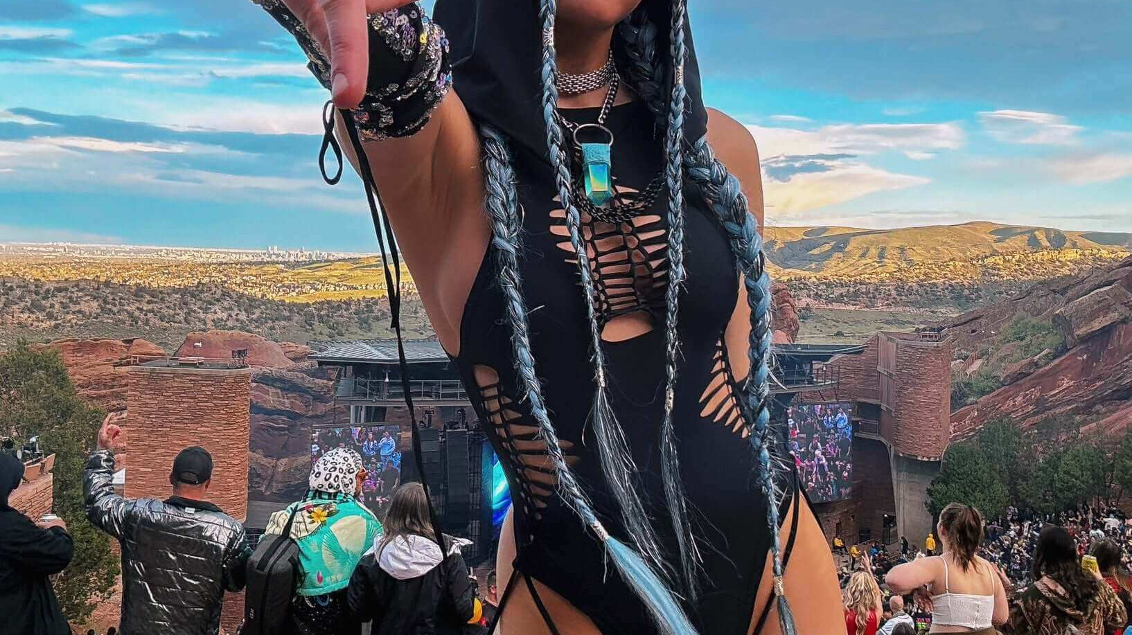 A festival-goer in a black rave bodysuit poses confidently with braided hair and accessories, set against a scenic outdoor venue
