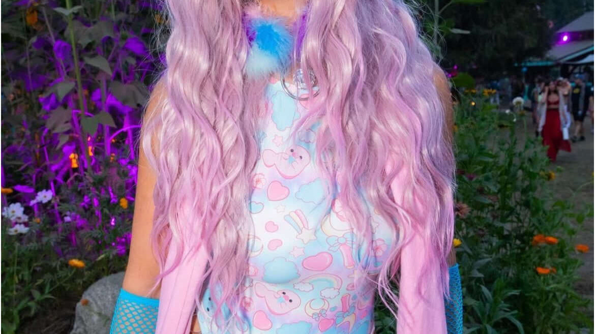 A festival-goer with long pink hair, blue sunglasses, and a pastel-themed bodysuit, standing in front of sunflowers and a colorful background.