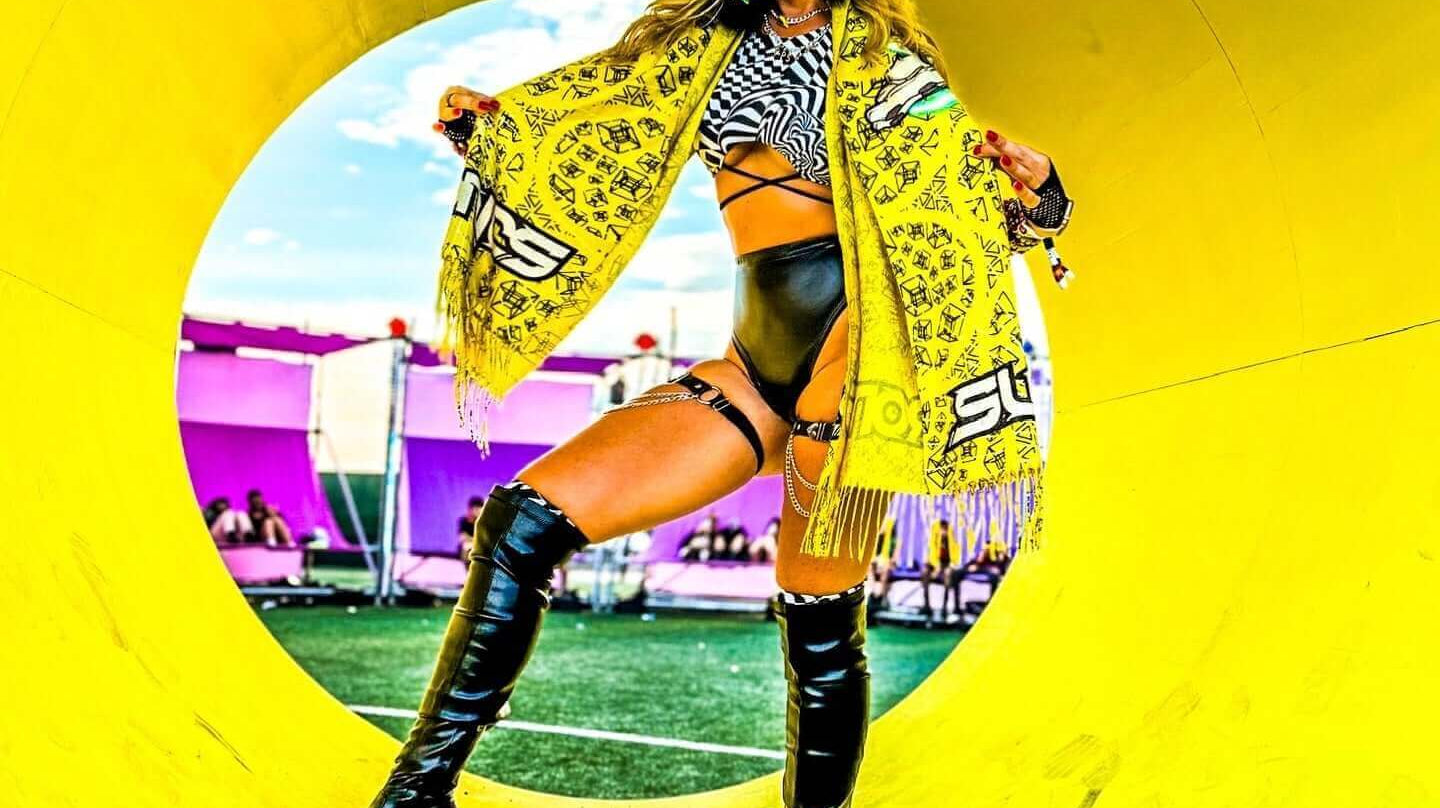 Woman in stylish rave outfit poses with a vibrant scarf in a colorful festival setting.
