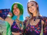Two women in floral Freedom Rave Wear Starflora outfits, one with green hair, under a clear blue sky