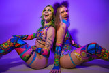 Two women sit back-to-back with playful expressions, in a room lit with purple lighting. They're wearing vibrant, stained-glass-inspired bodysuits and thigh-high stockings, and both have colorful makeup that complements their outfits. 