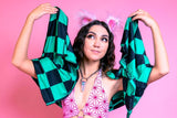 A woman with a whimsical expression holds up a green and black checkered pashmina by her head, wearing a pink patterned halter-neck top and fluffy pink ears, against a soft pink background. 