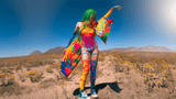 A colorful individual with vibrant green hair and a rainbow-hued outfit dances joyfully in a desert blooming with wildflowers, with mountainous terrain stretching into the distance under a clear blue sky.