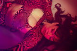 Woman in a swirl-patterned Freedom Rave Wear outfit with cutouts, under red-tinted light, in a dreamy pose.