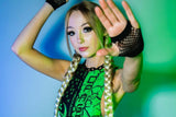 A woman with blonde braids raises her hand in front of her face, posing in a green and black rave outfit. The fishnet gloves and vibrant backdrop add a dynamic, modern feel to the image.