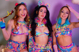 Three women sit closely together, smiling in front of a multicolored backdrop. They're wearing colorful, patterned rave outfits with cut-out details.
