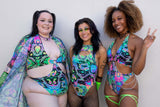  Three women stand together. They wear vibrant outfits with psychedelic prints; the first has a sheer cover-up, the second a cut-out rave bodysuit, and the third sports a halter-neck bodysuit. They celebrate diversity and style against a neutral backdrop.