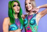 Two women pose against a lavender backdrop; one with green hair blowing a kiss and the other with blonde hair smiling, wearing tops with vivid psychedelic patterns.