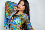 A woman with long dark hair poses with one hand near her face. She is wearing a psychedelic printed rave bodysuit.