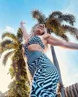  A joyous woman in a black and white patterned outfit reaching upwards, with palm trees and blue sky in the background.
