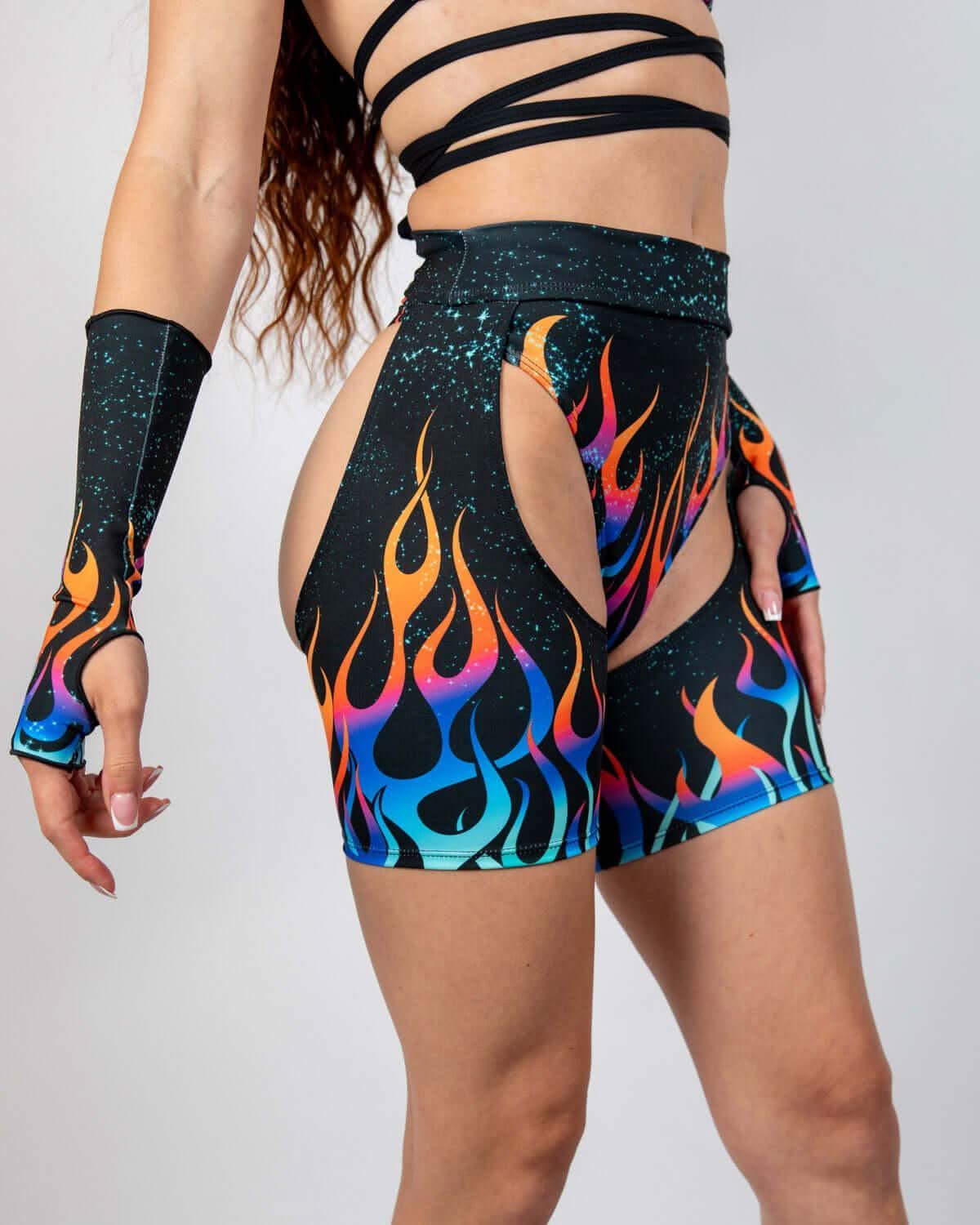 Model wearing Blaze Chaps in fiery flame graphics, showcasing high-waisted fit and open hip cutouts, ideal rave outfits.
