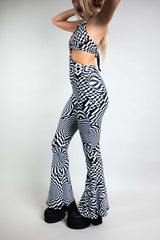 A woman wearing a black and white geometric printed jumpsuit with bell bottoms. The jumpsuit is a halter neck with cutouts on either side and an o-ring detail. The woman is facing to the left.