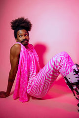 A man sitting in front of a pink backdrop wearing pink and white joggers with a matching tank top and a scarf draped over his shoulders.