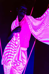 A man standing in black light, wearing a pink and white outfit with a matching pashmina draped over his shoulders. The pieces glow under the light.