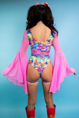 A woman wearing a rainbow bodysuit with whimsical characters printed on it, a lace up front detail, and flowy pink sleeves. She is facing away from the camera.