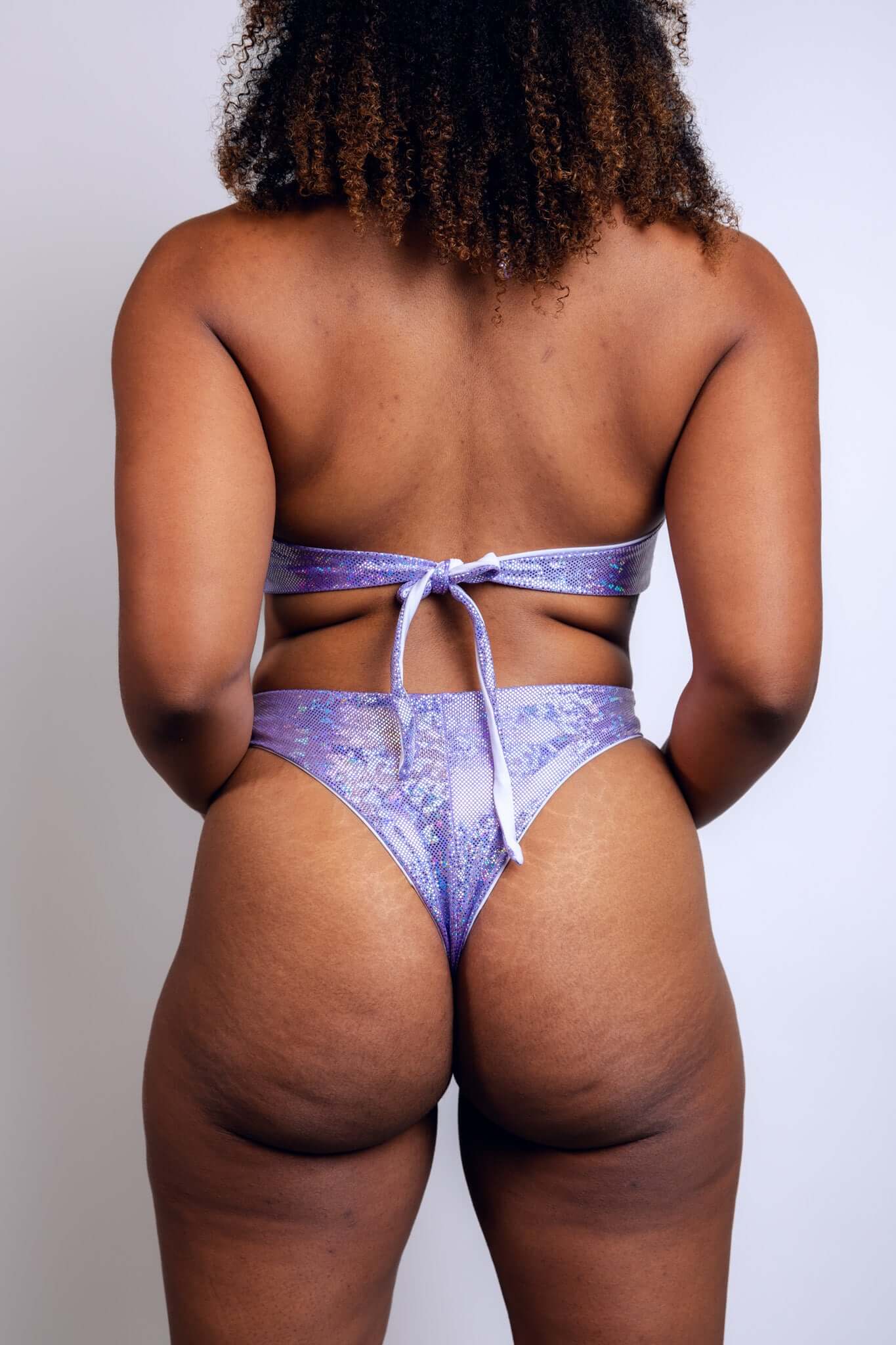 Rear view of a model in a lavender high-waisted bottom with a tie closure, showcasing the shimmering texture of the fabric from Freedom Rave Wear.