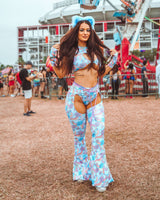 Rave enthusiast in a pastel blue festival outfit with floral patterns, fluffy ears, and bell-bottom rave pants at a bustling music event.