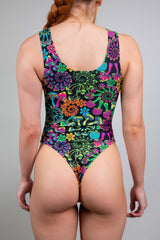 Rear view of a model in a Freedom Rave Wear Psybloom bodysuit, with a high neckline and vibrant neon floral pattern, set against a soft neutral backdrop.