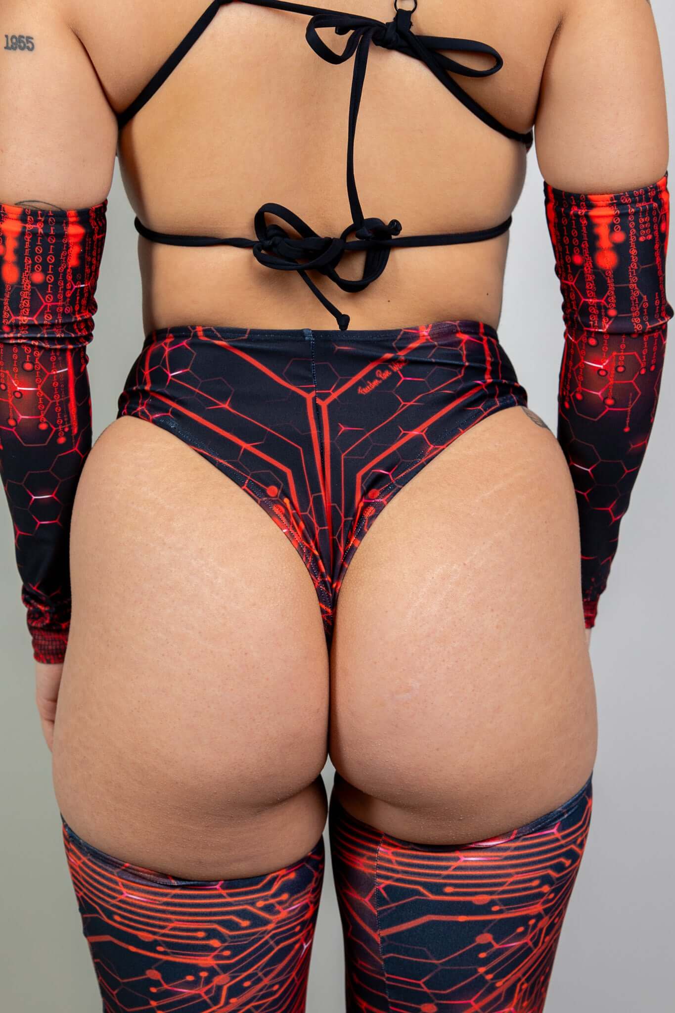 Rear view of a woman in a black and red Freedom Rave Wear high-waisted bottom featuring a circuit design, tied at the back, paired with matching arm sleeves