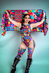 A girl with black swirly eye makeup holding up a rainbow patchwork pashmina behind her head. She is wearing a matching bodysuit.