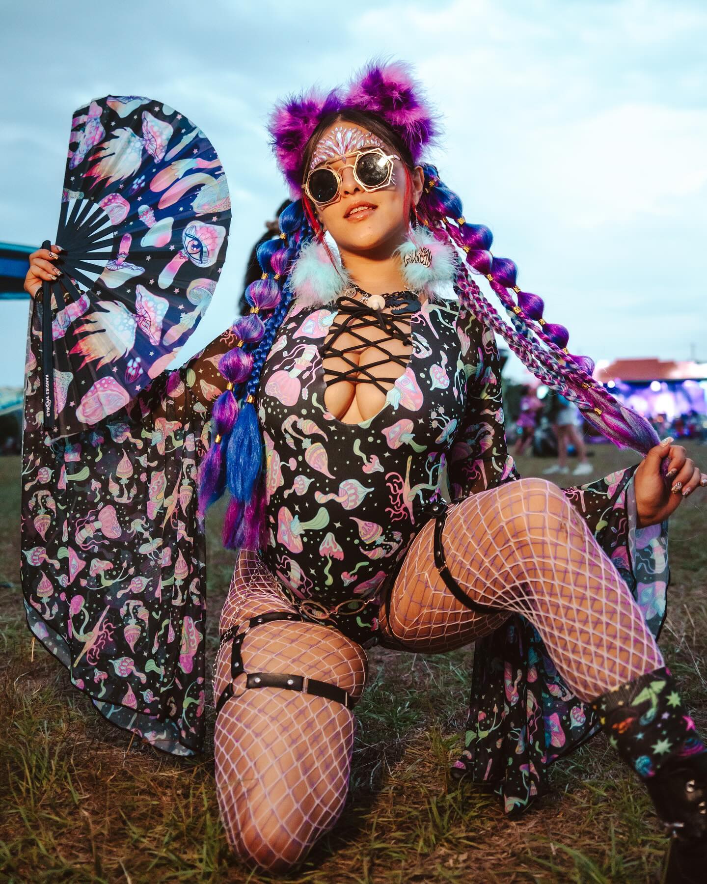 excited rave girl in a purple mushroom rave outfit with a matching fan and fluffy ear accessories and braids kneels in festival grounds