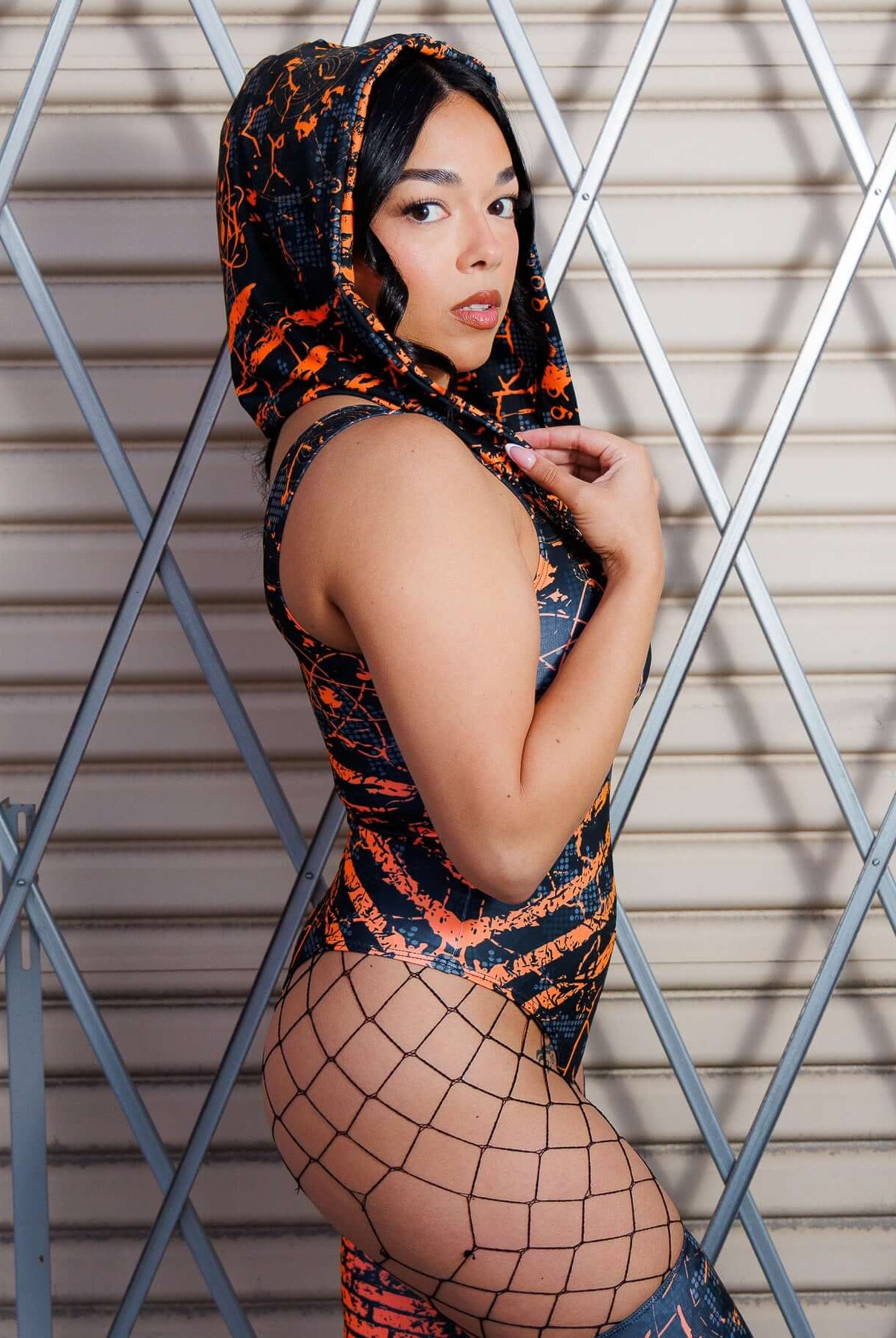 Model wearing Atomic Assassin Hood in orange and black, showcasing post-apocalyptic style with netted leggings against industrial backdrop.