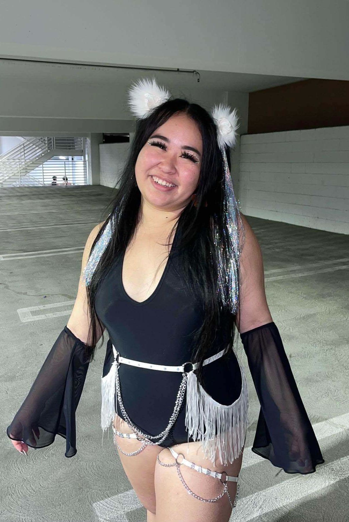 Young woman in a black bodysuit with black mesh flare sleeves, accessorized for a rave outfit, standing in a parking garage.