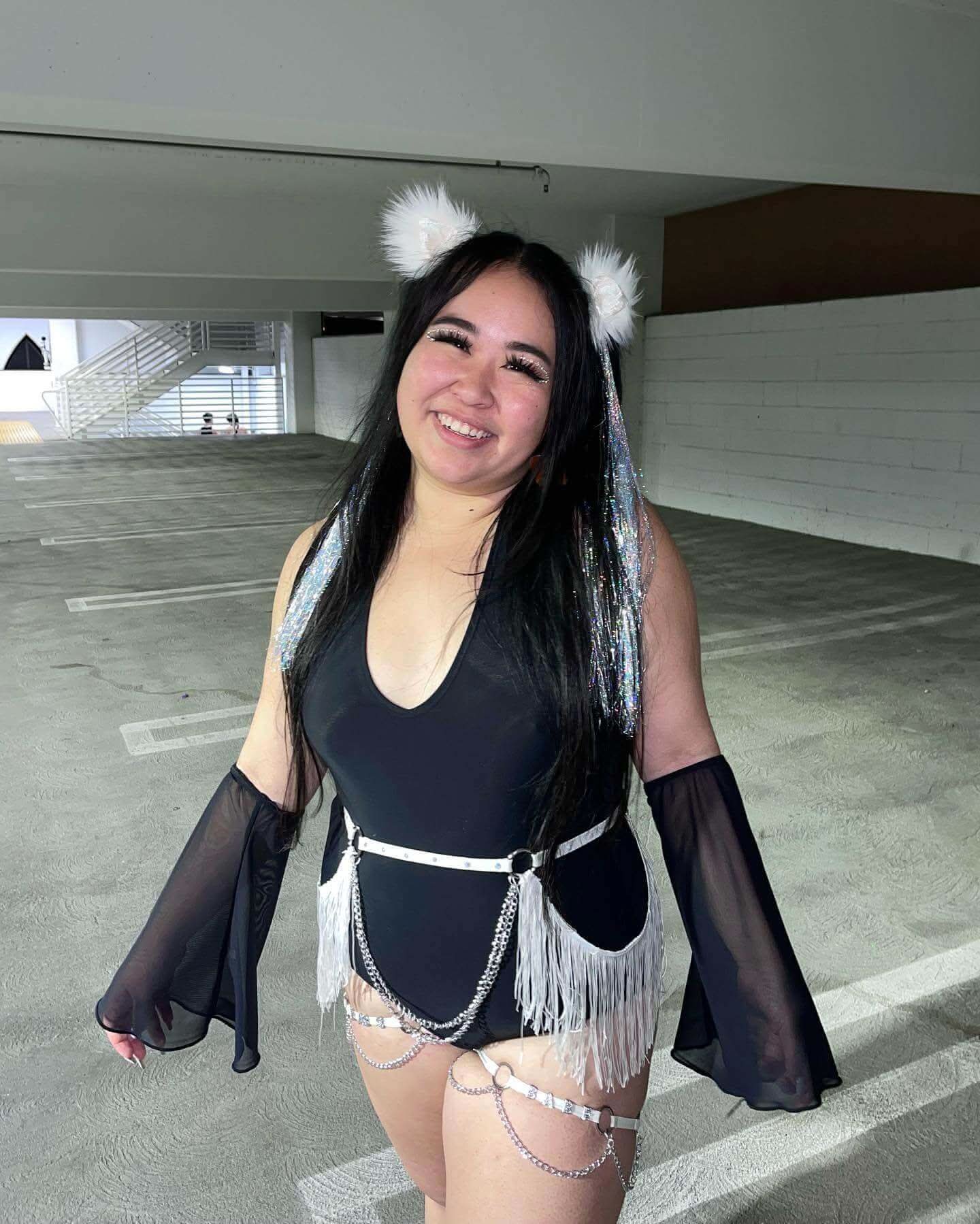 Young woman in a black bodysuit with black mesh flare sleeves, accessorized for a rave outfit, standing in a parking garage.