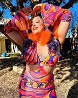 Radiant raver forms a heart with her hands, wearing a colorful outfit with a fluffy orange collar, basking in the sun’s glow