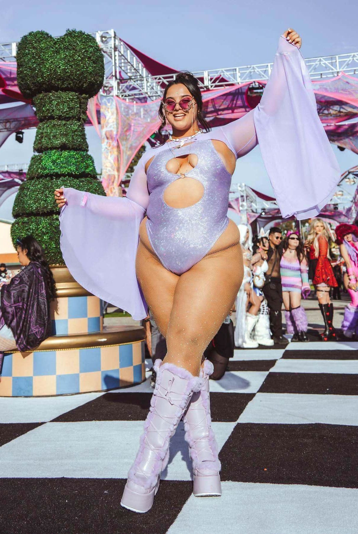 Model in lavender holo keyhole bodysuit with flowing sleeves, posing at a lively outdoor rave event.