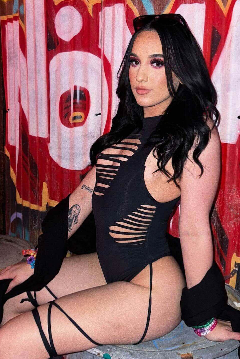 Model in a matte black slit sideboob bodysuit with leg straps, posing against graffiti, perfect for rave outfits.