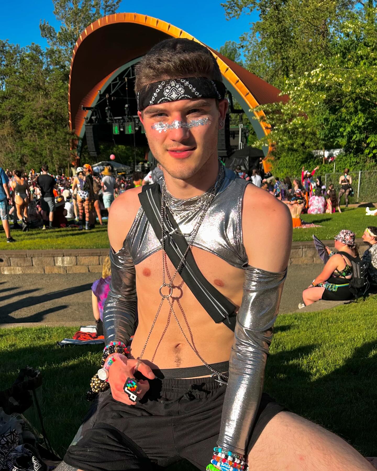 male raver in chrome metallic rave outfit with cutouts and glitter on face sits in front of stage in the festival crowd