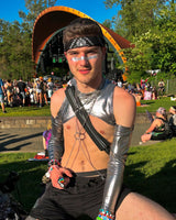 male raver in chrome metallic rave outfit with cutouts and glitter on face sits in front of stage in the festival crowd