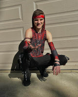 A smiling young man in a red and black rave outfit with bandana and arm sleeves kneels, offering a playful hand forward.