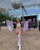 Joyful festival attendee dances under a giant disco ball, flaunting her vibrant, colorful rave outfit complete with floral patterns and matching knee-high leg sleeves, exuding a lively festival vibe.