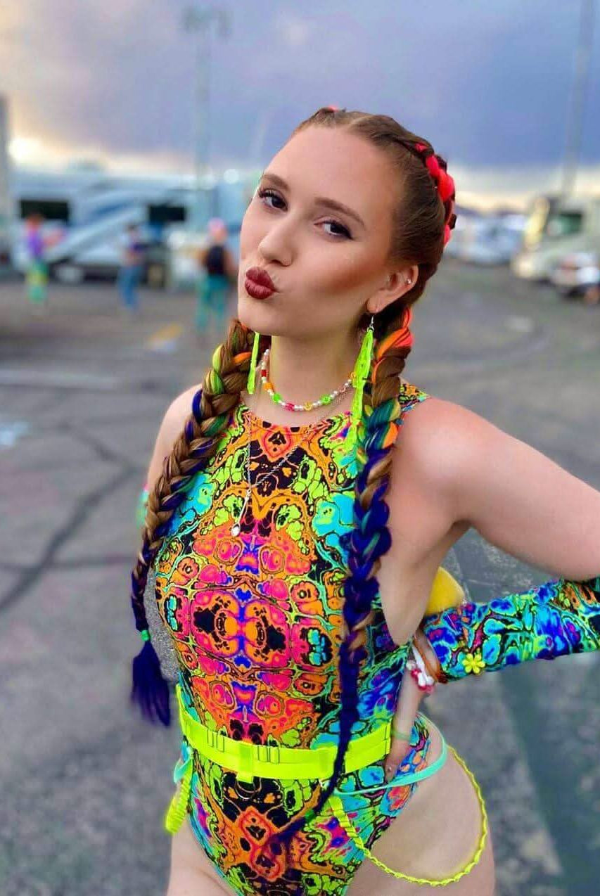 Colorful woman posing in a neon high-neck bodysuit, showcasing vibrant psychedelic print and bold accessories at a festival.