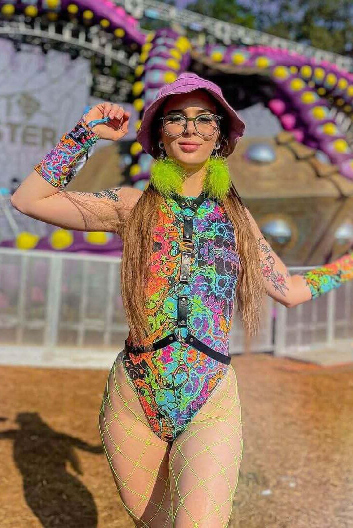 Model wearing a colorful Lucidity Sideboob Bodysuit at a rave festival, showcasing psychedelic rainbow print and vibrant accessories.