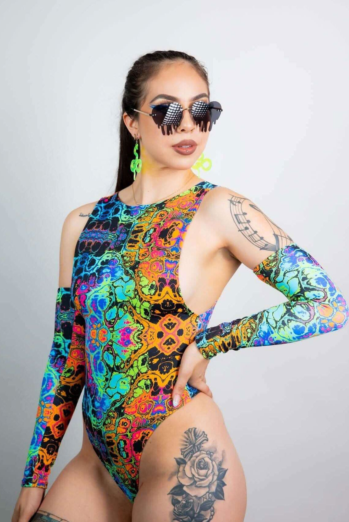Model wearing Lucidity Sideboob Bodysuit in psychedelic rainbow print, showcasing rave outfits style with sunglasses and accessories.